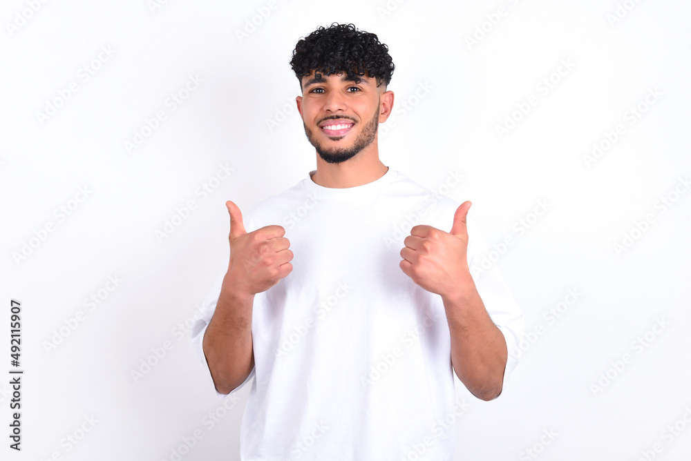 Optimistic young arab man with curly hair wearing white t-shirt over white background showing thumbs up with positive emotions. Quality and recommendation concept. Optimistic young arab man with curly hair wearing white t-shirt over white background showing thumbs up with positive emotions. Quality and recommendation concept.