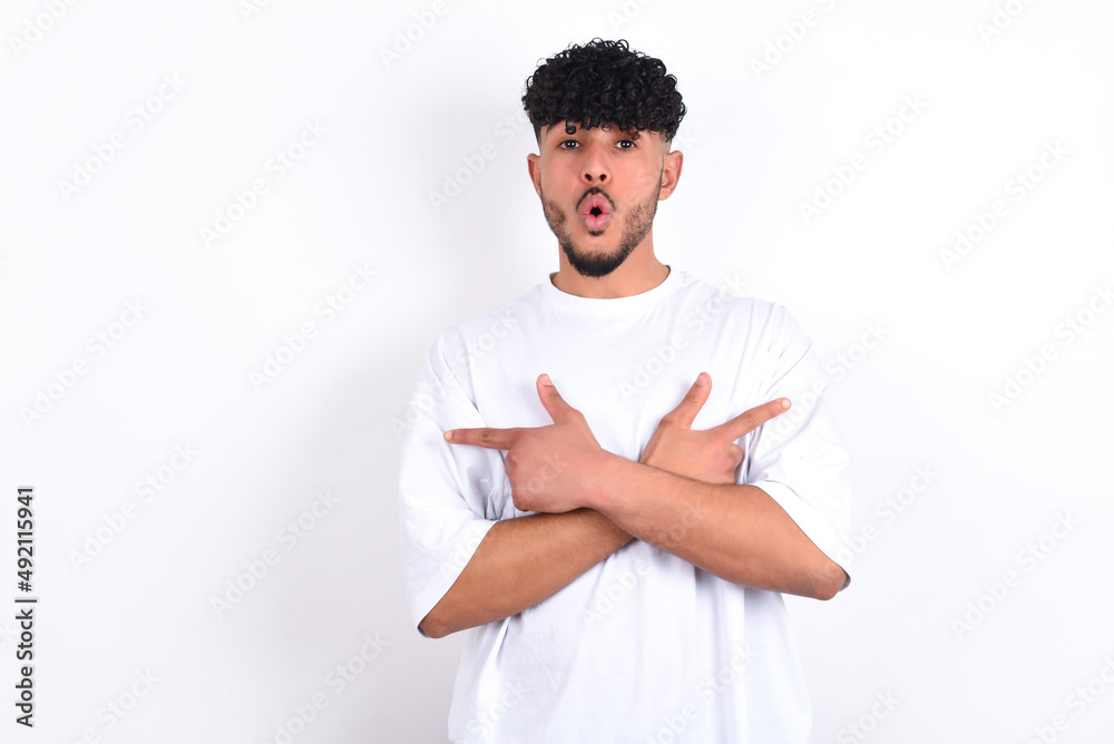 Confused young arab man with curly hair wearing white t-shirt over white background  chooses between two ways, points at both sides with crossed hands, feels doubt. Need your advice.