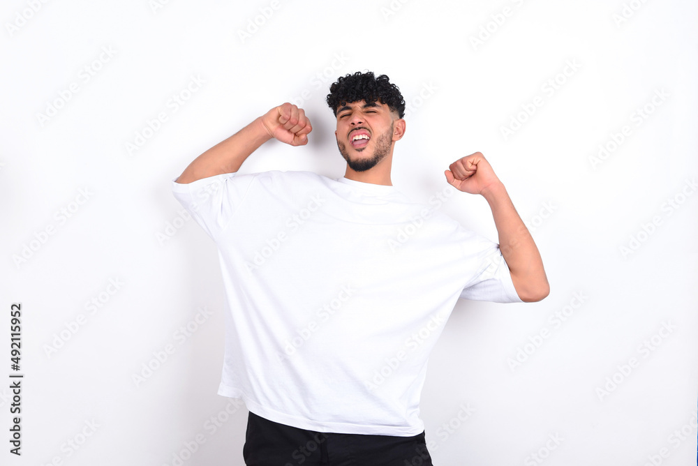 Yes I am winner. Portrait of charming delighted and excited young arab man with curly hair wearing white t-shirt raising up fist in triumph and victory smiling achieving success grinning from delight. Yes I am winner. Portrait of charming delighted and excited young arab man with curly hair wearing white t-shirt raising up fist in triumph and victory smiling achieving success grinning from delight.