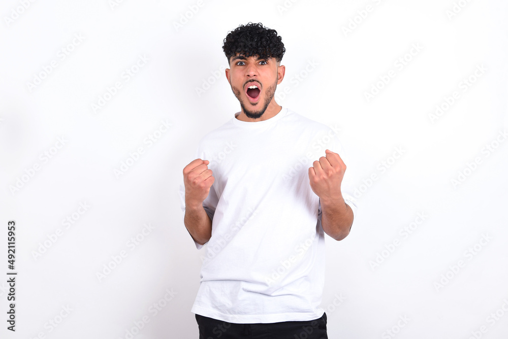 Joyful excited lucky young arab man with curly hair wearing white t-shirt over white background cheering, celebrating success, screaming yes with clenched fists Joyful excited lucky young arab man with curly hair wearing white t-shirt over white background cheering, celebrating success, screaming yes with clenched fists