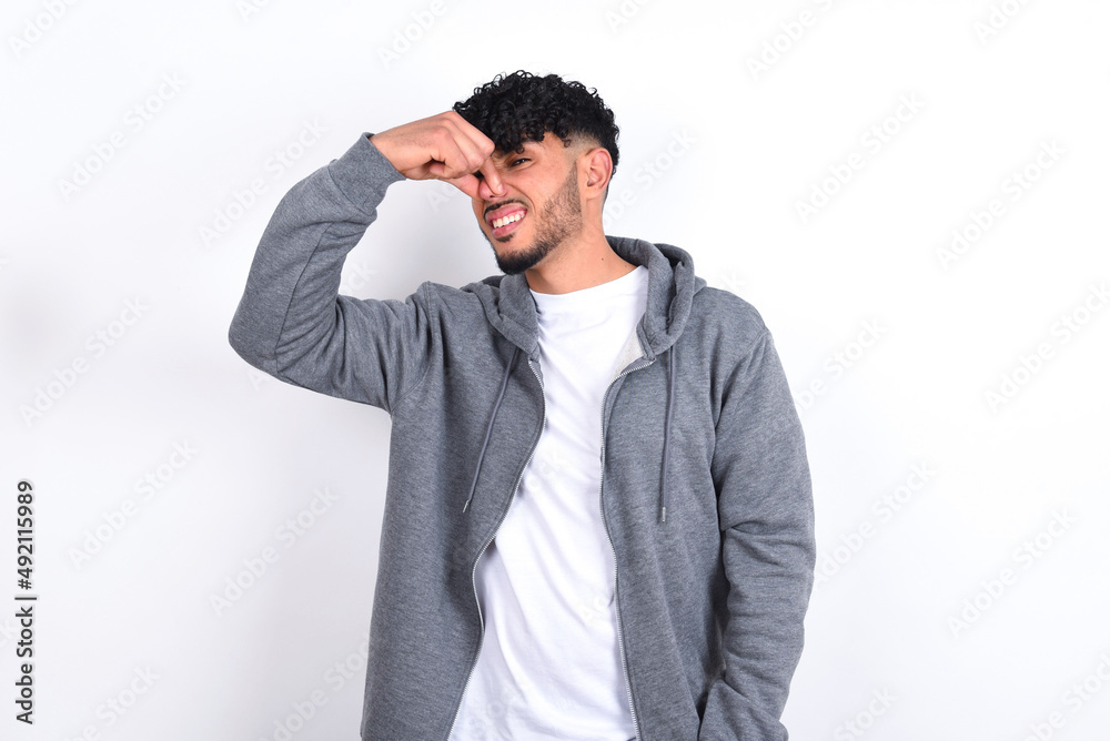 Displeased young arab man with curly hair wearing casual clothes over white background plugs nose as smells something stink and unpleasant, feels aversion, hates disgusting scent. Displeased young arab man with curly hair wearing casual clothes over white background plugs nose as smells something stink and unpleasant, feels aversion, hates disgusting scent.