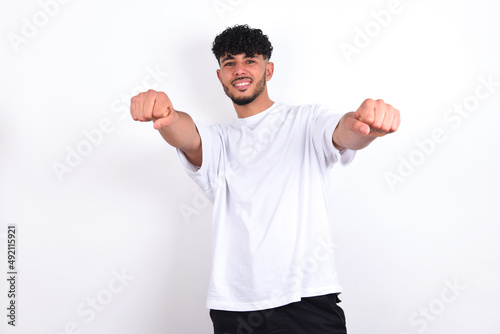 Portrait of charming young arab man with curly hair wearing white t-shirt over white background , smiling broadly while holding hands over her head. Confidence and relax concept.