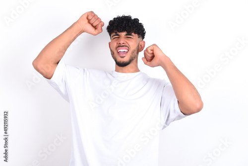 Attractive young arab man with curly hair wearing white t-shirt over white background celebrating a victory punching the air with his fists and a beaming toothy smile.