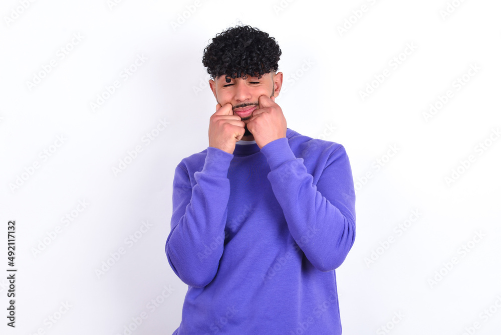 Pleased young arab man with curly hair wearing purple sweatshirt over white background with closed eyes keeps hands near cheeks and smiles tenderly imagines something very pleasant Pleased young arab man with curly hair wearing purple sweatshirt over white background with closed eyes keeps hands near cheeks and smiles tenderly imagines something very pleasant
