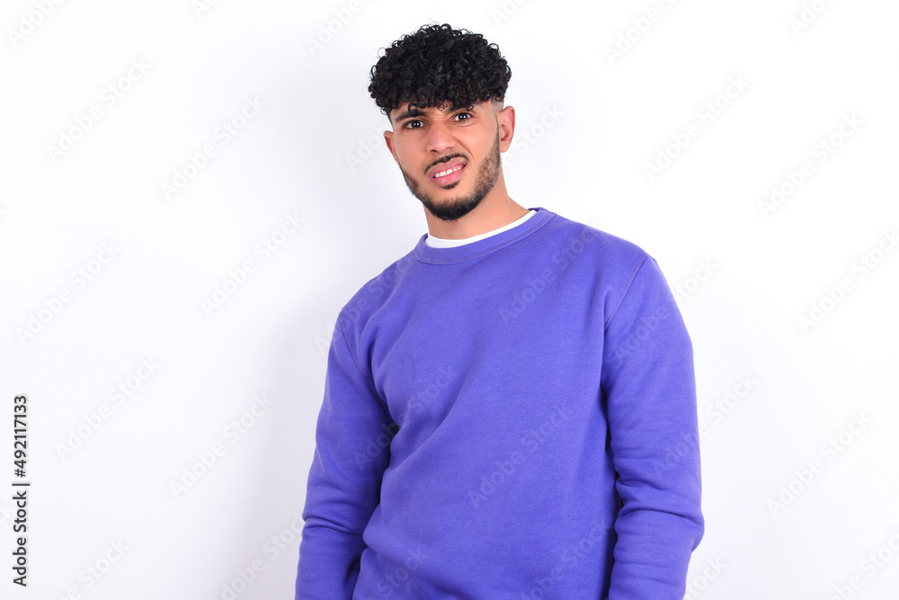 Portrait of dissatisfied young arab man with curly hair wearing purple sweatshirt over white wall smirks face, purses lips and looks with annoyance at camera, discontent hearing something unpleasant Portrait of dissatisfied young arab man with curly hair wearing purple sweatshirt over white wall smirks face, purses lips and looks with annoyance at camera, discontent hearing something unpleasant