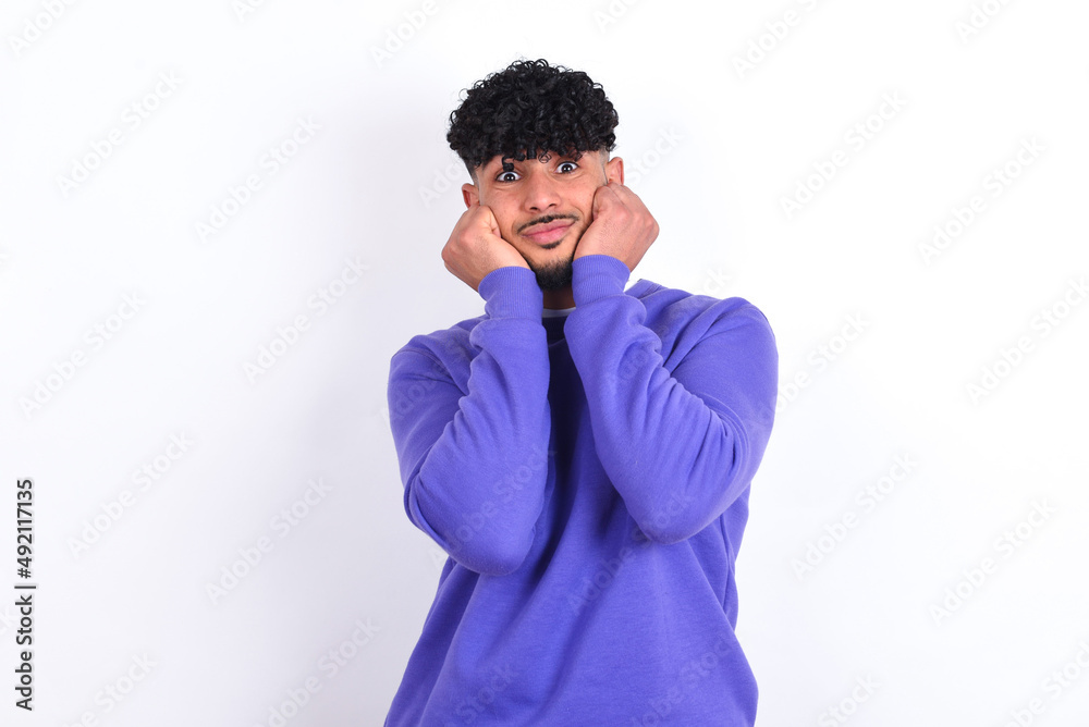 young arab man with curly hair wearing purple sweatshirt over white background with surprised expression keeps hands under chin keeps lips folded makes funny grimace young arab man with curly hair wearing purple sweatshirt over white background with surprised expression keeps hands under chin keeps lips folded makes funny grimace