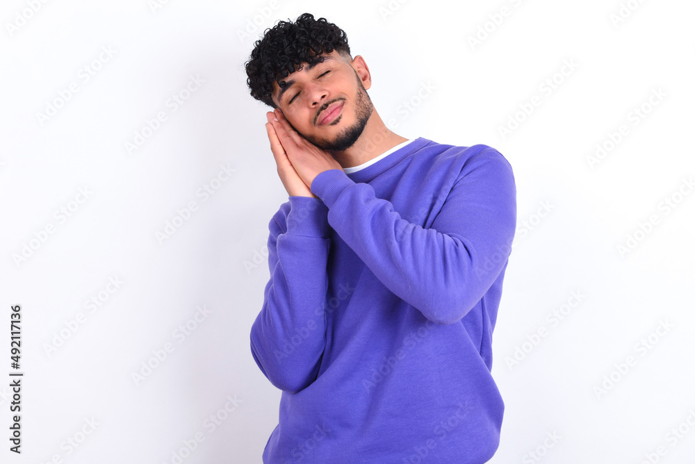young arab man with curly hair wearing purple sweatshirt over white background leans on pressed palms closes eyes and has pleasant smile dreams about something young arab man with curly hair wearing purple sweatshirt over white background leans on pressed palms closes eyes and has pleasant smile dreams about something