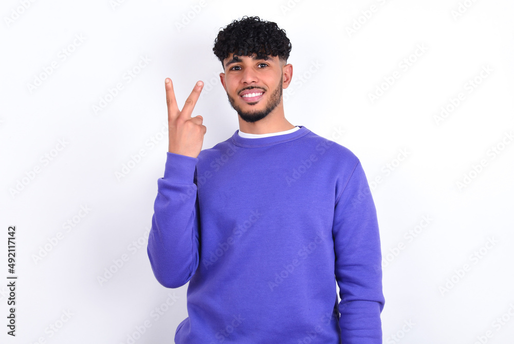 young arab man with curly hair wearing purple sweatshirt over white background smiling and looking friendly, showing number two or second with hand forward, counting down young arab man with curly hair wearing purple sweatshirt over white background smiling and looking friendly, showing number two or second with hand forward, counting down