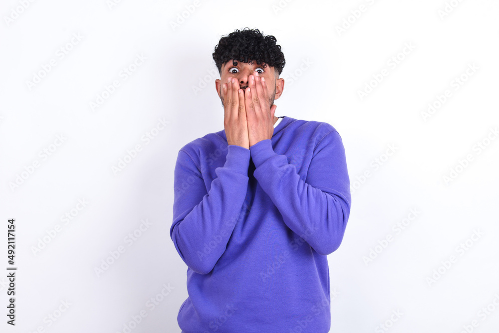young arab man with curly hair wearing purple sweatshirt over white background keeps hands on mouth, looks with eyes full of disbelief, being puzzled with amount of work young arab man with curly hair wearing purple sweatshirt over white background keeps hands on mouth, looks with eyes full of disbelief, being puzzled with amount of work