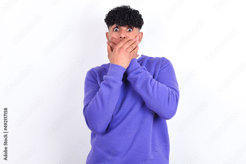 Stunned young arab man with curly hair wearing purple sweatshirt over white background covers mouth with both hands being afraid from something or after hearing stunning gossips. Stunned young arab man with curly hair wearing purple sweatshirt over white background covers mouth with both hands being afraid from something or after hearing stunning gossips.
