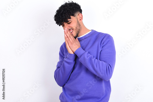 Overjoyed young arab man with curly hair wearing purple sweatshirt over white background laughs joyfully and keeps palms pressed together hears something funny