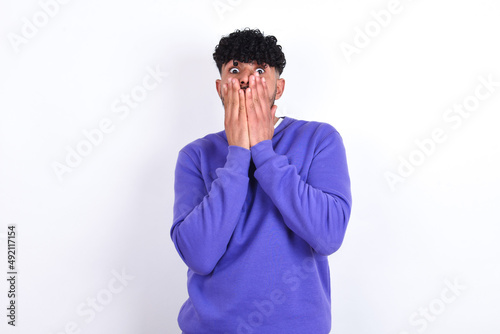 young arab man with curly hair wearing purple sweatshirt over white background keeps hands on mouth, looks with eyes full of disbelief, being puzzled with amount of work