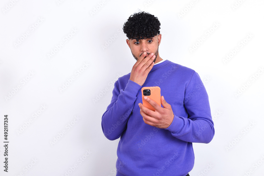 young arab man with curly hair wearing purple sweatshirt over white background being deeply surprised, stares at smartphone display, reads shocking news on website, Omg, its horrible! young arab man with curly hair wearing purple sweatshirt over white background being deeply surprised, stares at smartphone display, reads shocking news on website, Omg, its horrible!