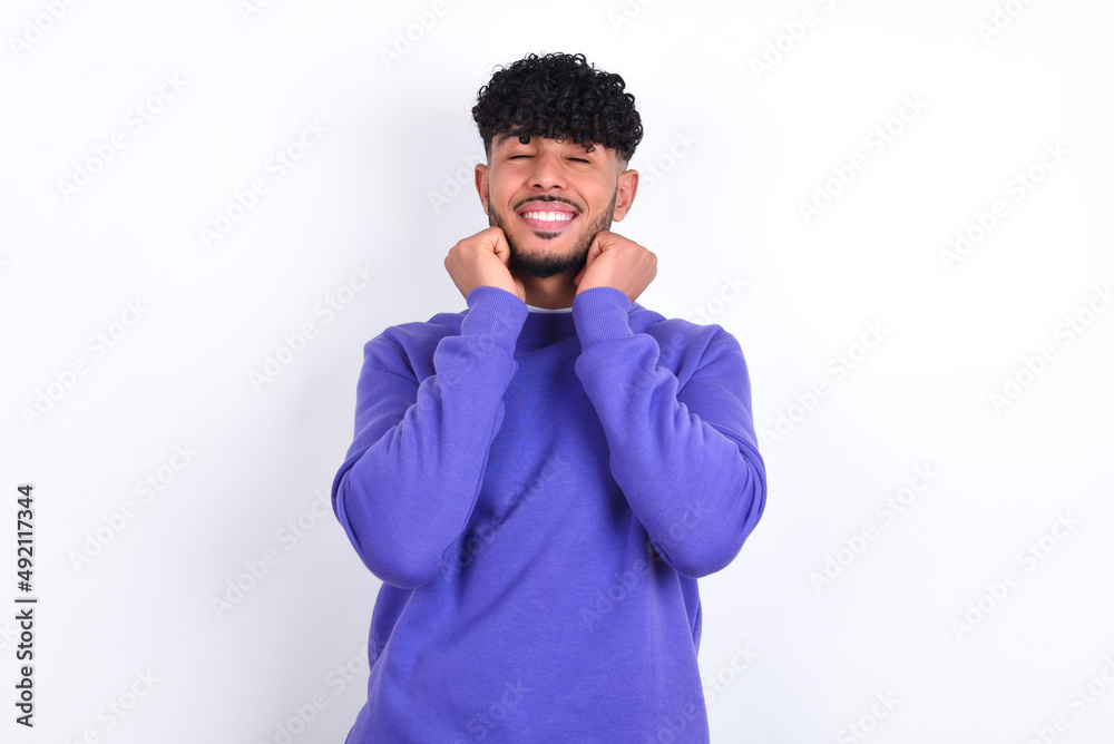 young arab man with curly hair wearing purple sweatshirt over white background grins joyfully, imagines something pleasant, copy space. Pleasant emotions concept. young arab man with curly hair wearing purple sweatshirt over white background grins joyfully, imagines something pleasant, copy space. Pleasant emotions concept.