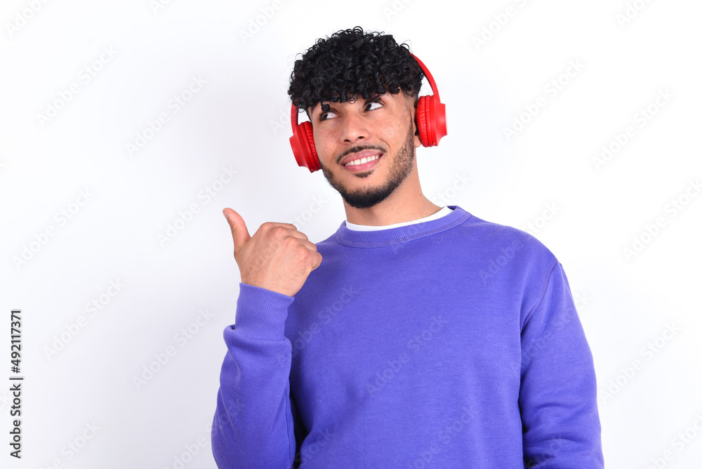 young arab man with curly hair wearing purple sweatshirt over white background listens audio track via wireless headphones points thumb away advertises copy space young arab man with curly hair wearing purple sweatshirt over white background listens audio track via wireless headphones points thumb away advertises copy space