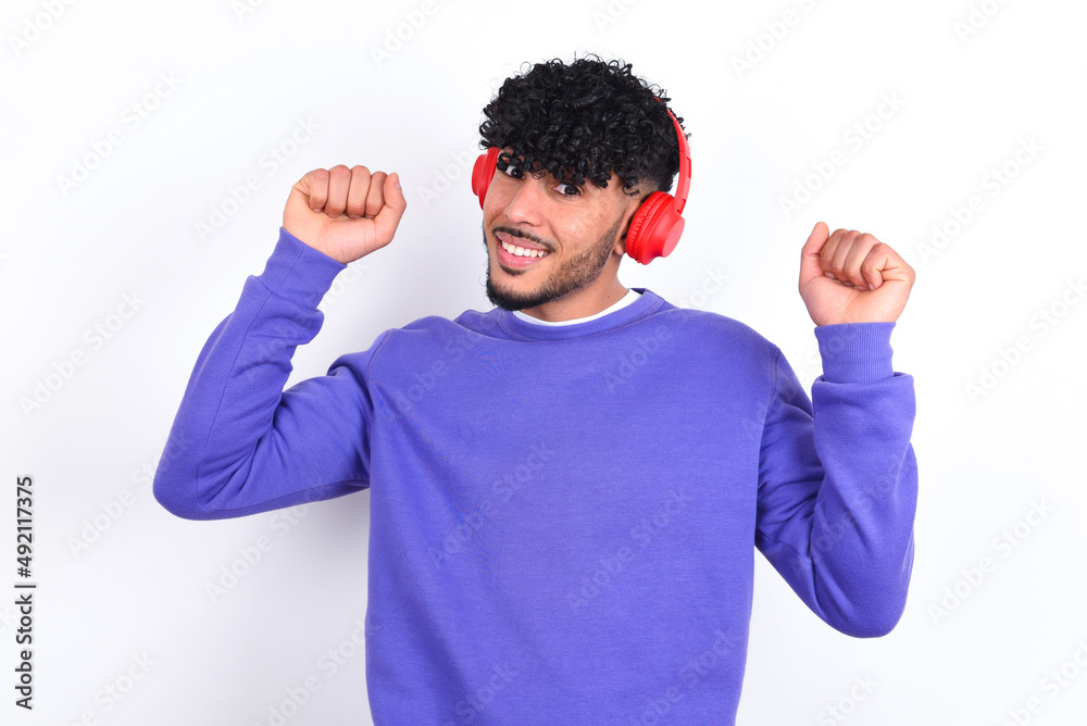 Carefree young arab man with curly hair wearing purple sweatshirt over white wall with toothy smile raises arms dances carefree moves with rhythm of music listens music from playlist via headphones Carefree young arab man with curly hair wearing purple sweatshirt over white wall with toothy smile raises arms dances carefree moves with rhythm of music listens music from playlist via headphones