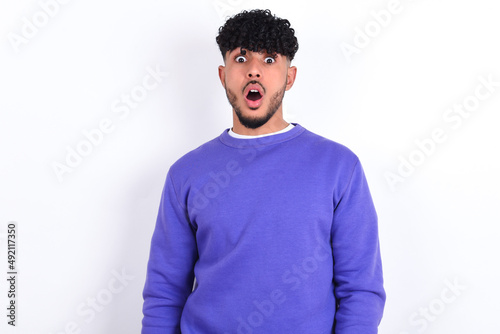 Shocked young arab man with curly hair wearing purple sweatshirt over white background stares bugged eyes keeps mouth opened has surprised expression. Omg concept