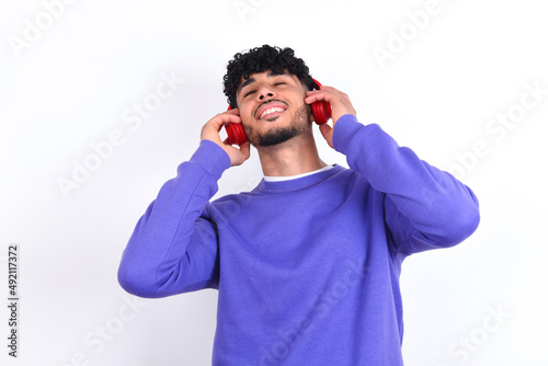 young arab man with curly hair wearing purple sweatshirt over white background smiles broadly feels very glad listens favourite music track via wireless headphones closes eyes.