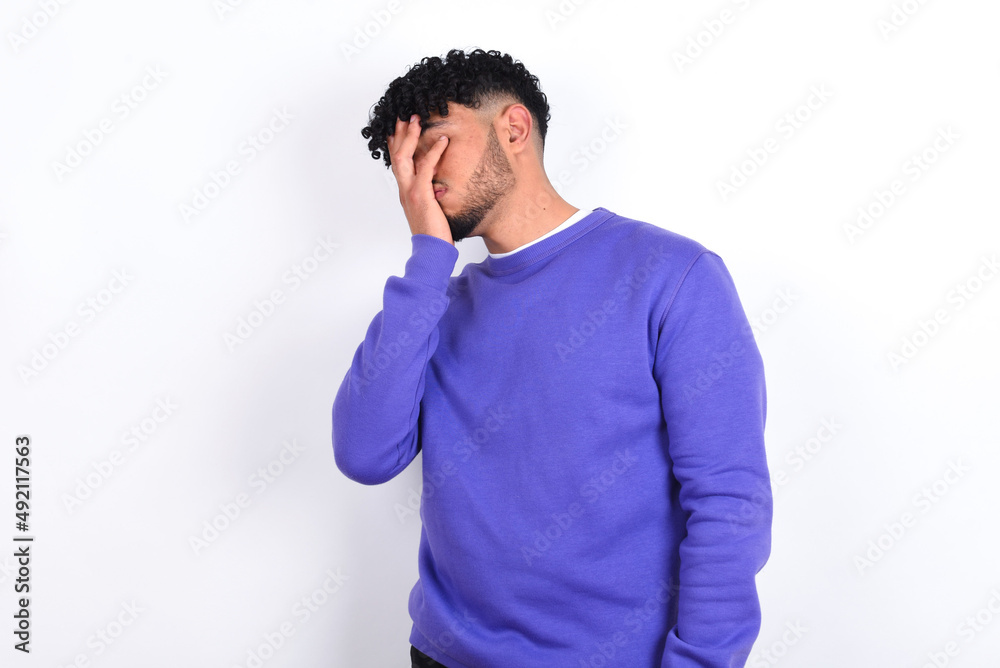 young arab man with curly hair wearing purple sweatshirt over white background with sad expression covering face with hands while crying. Depression concept. young arab man with curly hair wearing purple sweatshirt over white background with sad expression covering face with hands while crying. Depression concept.
