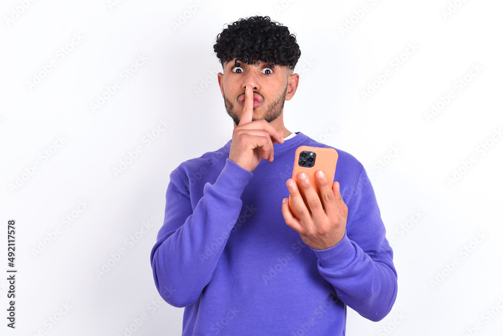 young arab man with curly hair wearing purple sweatshirt over white background holding modern gadget ask not tell secrets young arab man with curly hair wearing purple sweatshirt over white background holding modern gadget ask not tell secrets