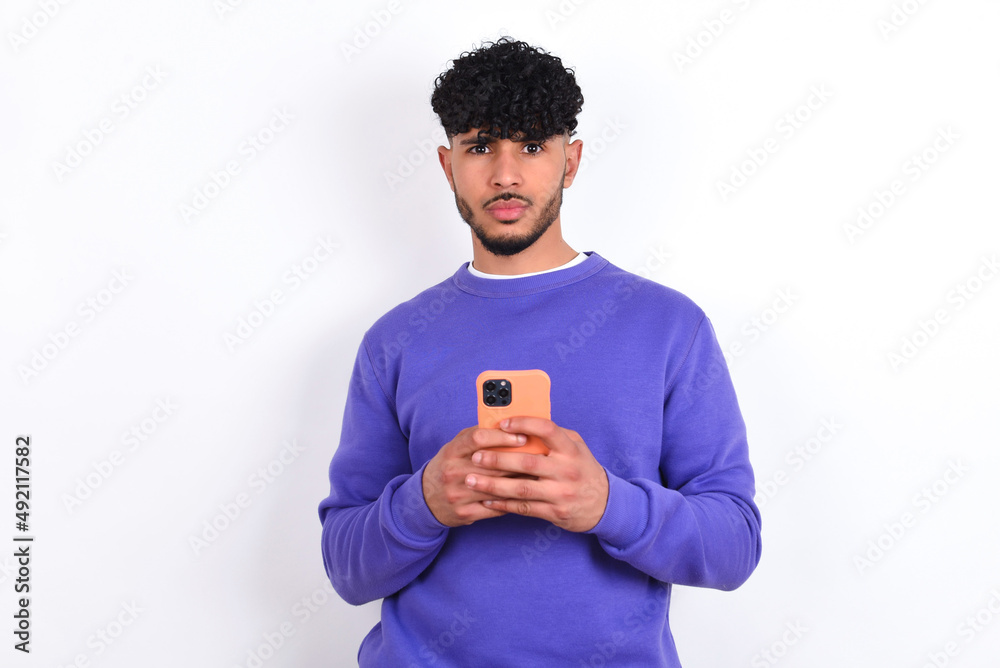 Portrait of serious confident young arab man with curly hair wearing purple sweatshirt over white background holding phone in two hands Portrait of serious confident young arab man with curly hair wearing purple sweatshirt over white background holding phone in two hands