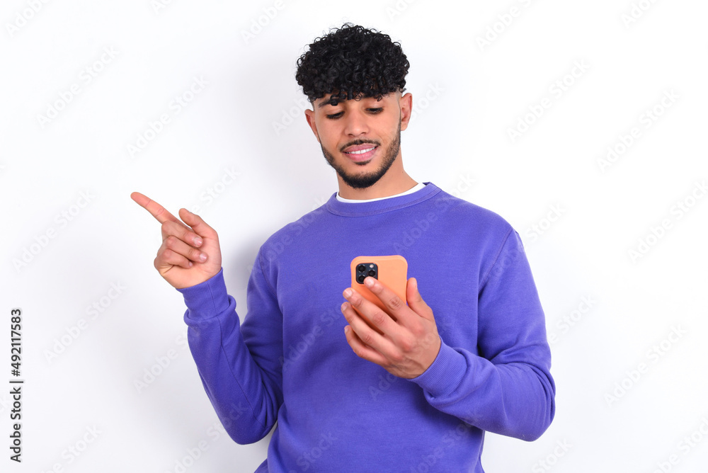 Smiling young arab man with curly hair wearing purple sweatshirt over white background pointing finger at blank space holding phone in one hand Smiling young arab man with curly hair wearing purple sweatshirt over white background pointing finger at blank space holding phone in one hand