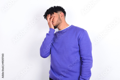 young arab man with curly hair wearing purple sweatshirt over white background with sad expression covering face with hands while crying. Depression concept.
