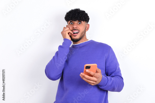 Afraid funny young arab man with curly hair wearing purple sweatshirt over white background holding telephone and bitting nails