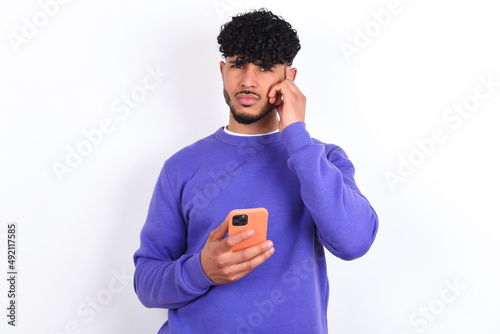 young arab man with curly hair wearing purple sweatshirt over white background holding gadget while sticking out tongue