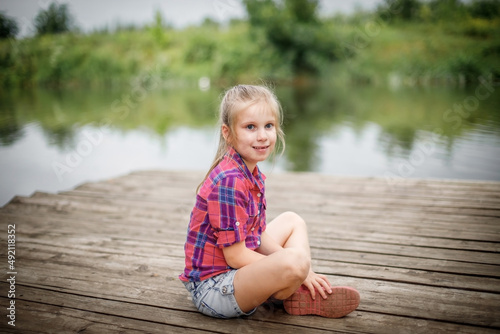 A blonde girl sits on a wooden pier by the river.