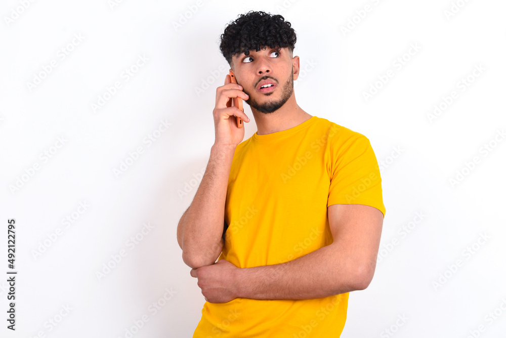 Sad young arab man wearing yellow T-shirt over white background talking on smartphone. Communication concept. Sad young arab man wearing yellow T-shirt over white background talking on smartphone. Communication concept.