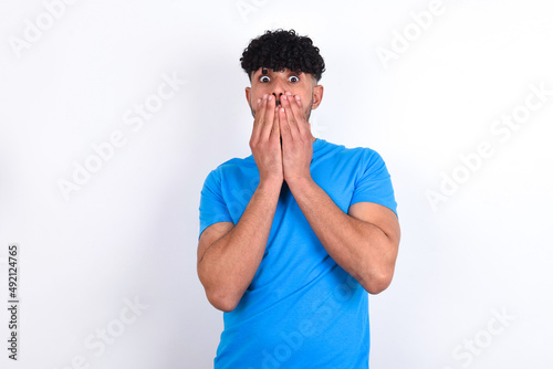 young arab man with curly hair wearing blue t-shirt over white background keeps hands on mouth, looks with eyes full of disbelief, being puzzled with amount of work