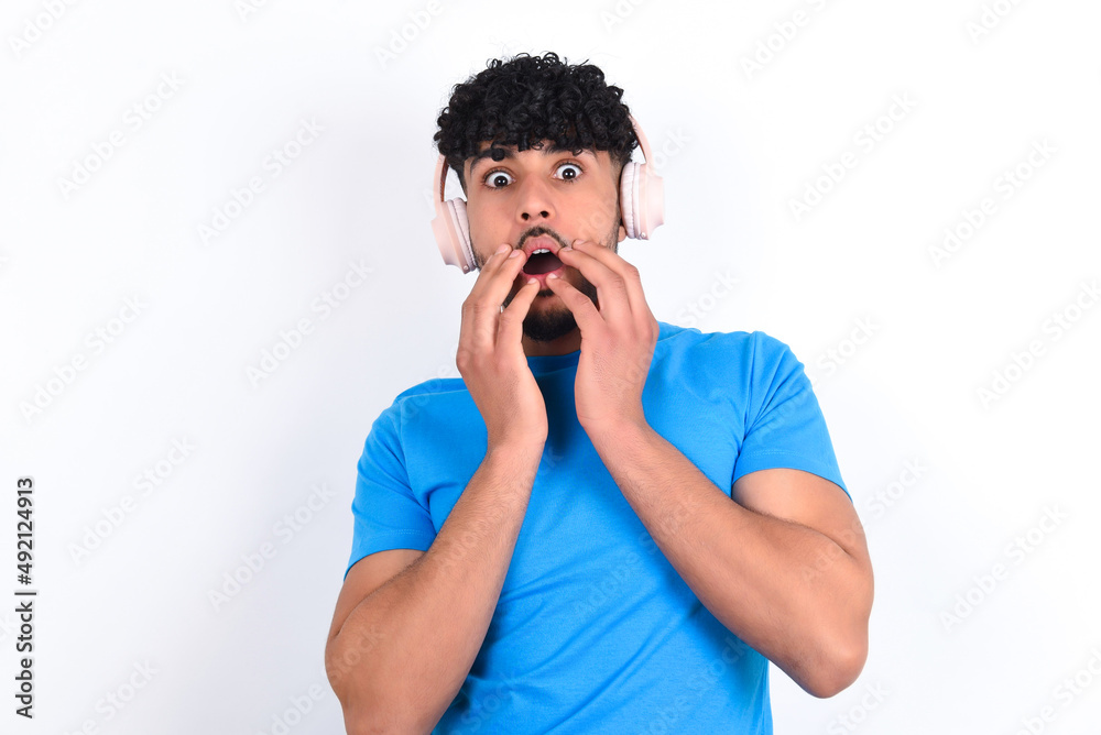 Shocked young arab man with curly hair wearing blue t-shirt over white background stares fearful at camera keeps mouth widely opened wears wireless stereo headphones on ears Shocked young arab man with curly hair wearing blue t-shirt over white background stares fearful at camera keeps mouth widely opened wears wireless stereo headphones on ears