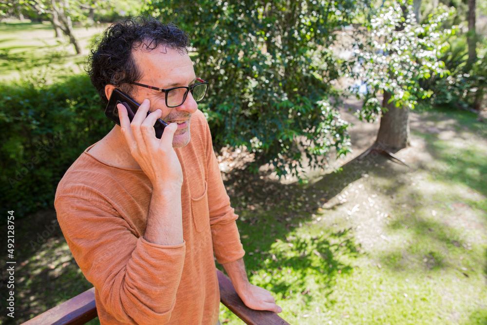 Entrepreneur man with glasses doing home office work with a smartphone on the balcony surrounding by trees during springtime. Self employer concept. Copy space. Entrepreneur man with glasses doing home office work with a smartphone on the balcony surrounding by trees during springtime. Self employer concept. Copy space.