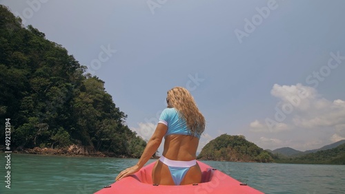 Sexy young woman in blue swimsuit swings feet in water on pink canoe on sea against hills backside view. Traveling to tropical countries. Attractive sportive girl is sailing on kayak in ocean. photo