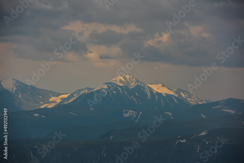 Awesome aerial view to high snow mountain peak in sunlight in overcast. Atmospheric mountain landscape at high altitude with cloudiness. Alpine view to snowy mountain top in light under cloudy sky.