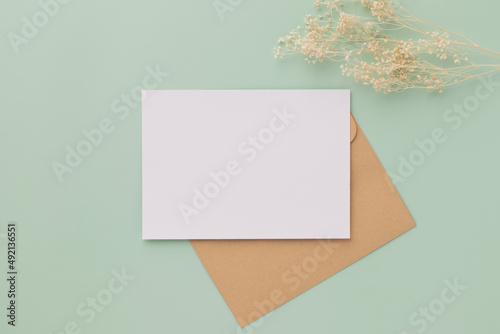 Blank greeting card invitation Mockup 5x7 on Brown envelope with dried flowers on pastel green background, flat lay, mockup photo