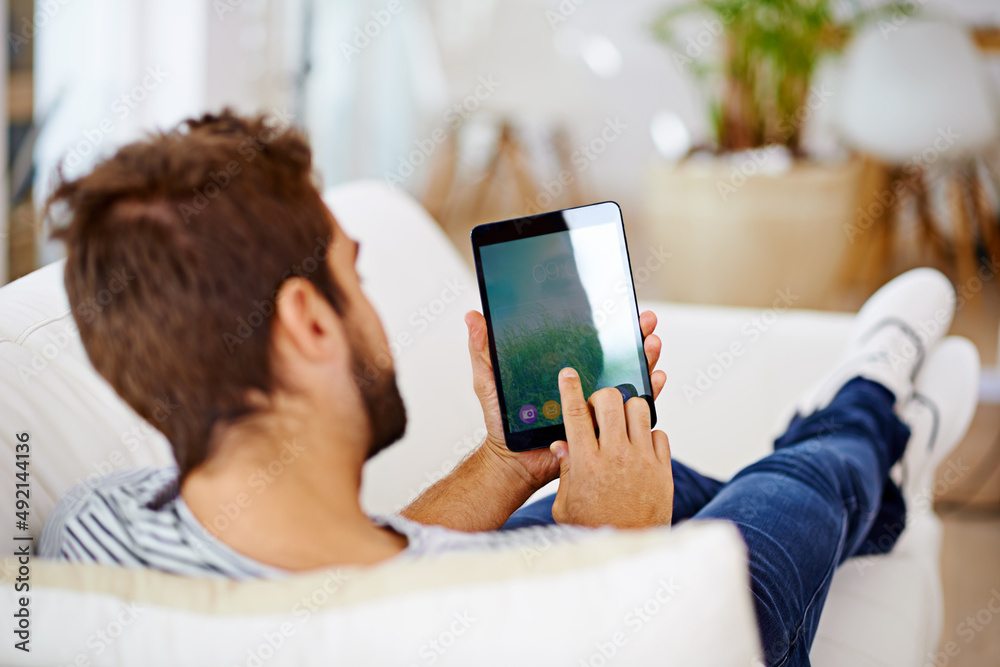 Enjoying some down-time with his latest tech. Rearview of a man using his tablet on his couch. Enjoying some down-time with his latest tech. Rearview of a man using his tablet on his couch.