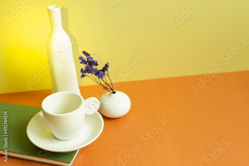 Notebook, white coffee cup, vase of purple dry flower on orange table. yellow wall background