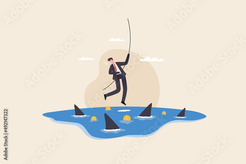 businessman hangs on for dear life from a rope as he swings above a group of circling sharks in the ocean. Risk taker, challenge to success, overcome difficulty or problem in crisis.