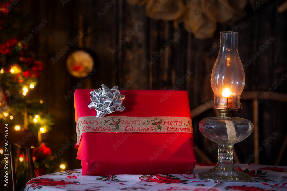 A Christmas gift wrapped in red paper and bow beside a lit retro oil lamp on a table top