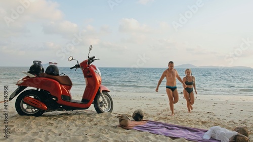 Fototapeta Naklejka Na Ścianę i Meble -  Scooter road trip. Lovely couple on red motorbike in white clothes on sand beach. Just married people kiss hugs walking near the tropical palm trees, sea. Wedding honeymoon by ocean. Motorcycle rent.