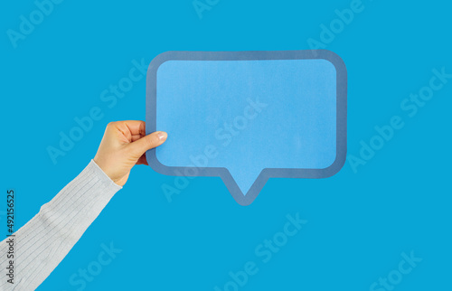 Human hand holding blank blue text message bubble. Young female showing rectangular paper mockup speech balloon. Cropped close up studio shot on blue background. Anonymous opinion concept