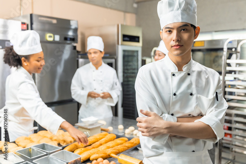 Smiling bakers looking at camera..Chef  baker in a chef dress and hat, cooking together in kitchen.Young asian man takes fresh baked cookies out of modern electric oven in kitchen. photo