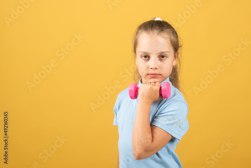 Child girl is doing exercises with dumbbells on yellow studio background.