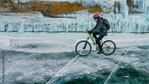 Man is riding bicycle near ice grotto. Rock with ice caves icicles. Teenage is dressed in black down jacket, cycling backpack, helmet. Tires on covered with special spikes. Traveler boy is ride cycle. © ivandanru