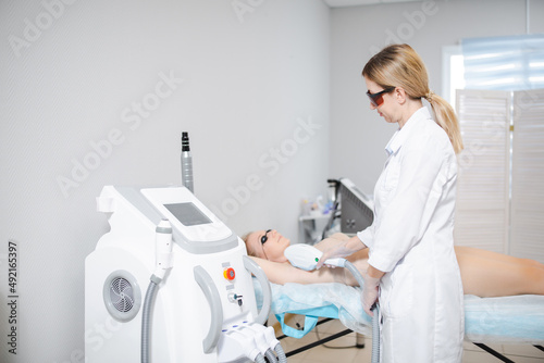 Photo of a modern cosmetology device and a female dermatologist in protective glasses performing a procedure of laser epilation of the armpits of a young girl