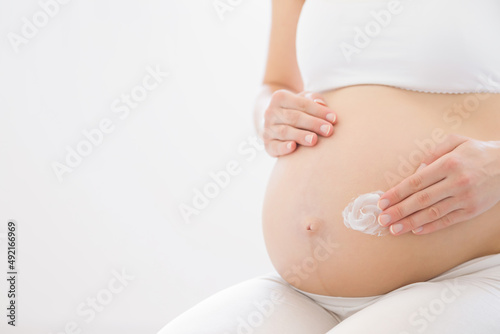 Young adult pregnant woman hand applying white moisturizer cream on big belly on light gray background. Care about perfect, soft and smooth body skin in pregnancy time. Closeup. Empty place for text. © fotoduets