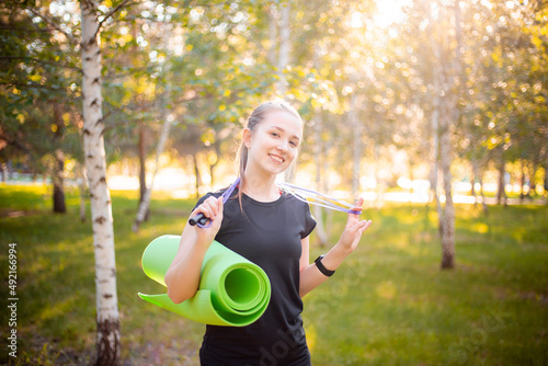 Portrait of a beautiful smiling young woman in a sports uniform with a jump rope and a yoga mat in her hands, posing against the backdrop of a park.