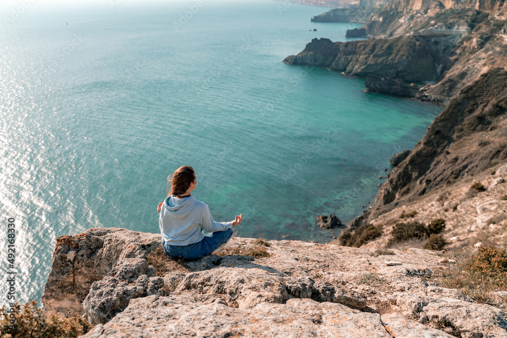 Woman tourist enjoying the sunset over the sea mountain landscape. Sits outdoors on a rock above the sea. She is wearing jeans and a blue hoodie. Healthy lifestyle, harmony and meditation Woman tourist enjoying the sunset over the sea mountain landscape. Sits outdoors on a rock above the sea. She is wearing jeans and a blue hoodie. Healthy lifestyle, harmony and meditation